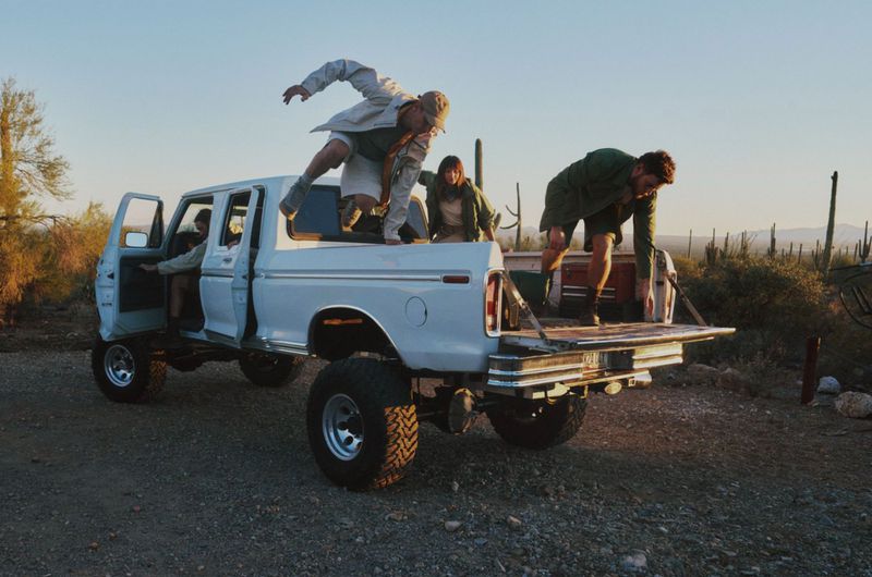 Four models interacting with a lifted white pickup truck in a desert landscape with cacti under a sunset sky