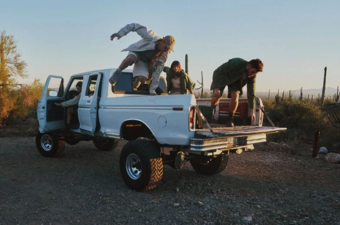 Four models interacting with a lifted white pickup truck in a desert landscape with cacti under a sunset sky