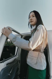 East Asian woman in a cream belted Moncler jacket and green pants standing beside an open car door against a pale sky.