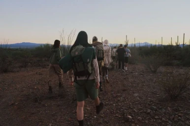 A group of hikers wearing backpacks walks along a trail through a desert landscape with saguaro cacti at twilight.