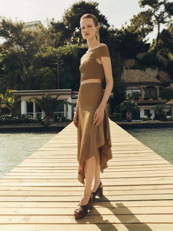 Model wearing a camel knit two-piece set and brown platform sandals poses on a wooden pier by the water for Elle Shoes Summer 26.