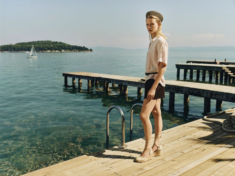 Model wearing espadrille wedge sandals, a short-sleeve shirt, and a hat standing on a wooden pier by the sea.