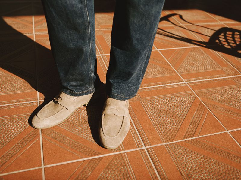 Close-up of a person wearing dark denim jeans and beige suede loafers standing on a terracotta patterned tile floor.