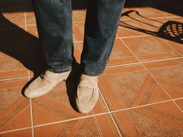 Close-up of a person wearing dark denim jeans and beige suede loafers standing on a terracotta patterned tile floor.