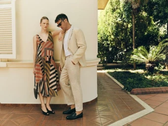A man in a beige linen suit and sunglasses standing beside a woman in a geometric pattern dress on a tiled villa terrace.