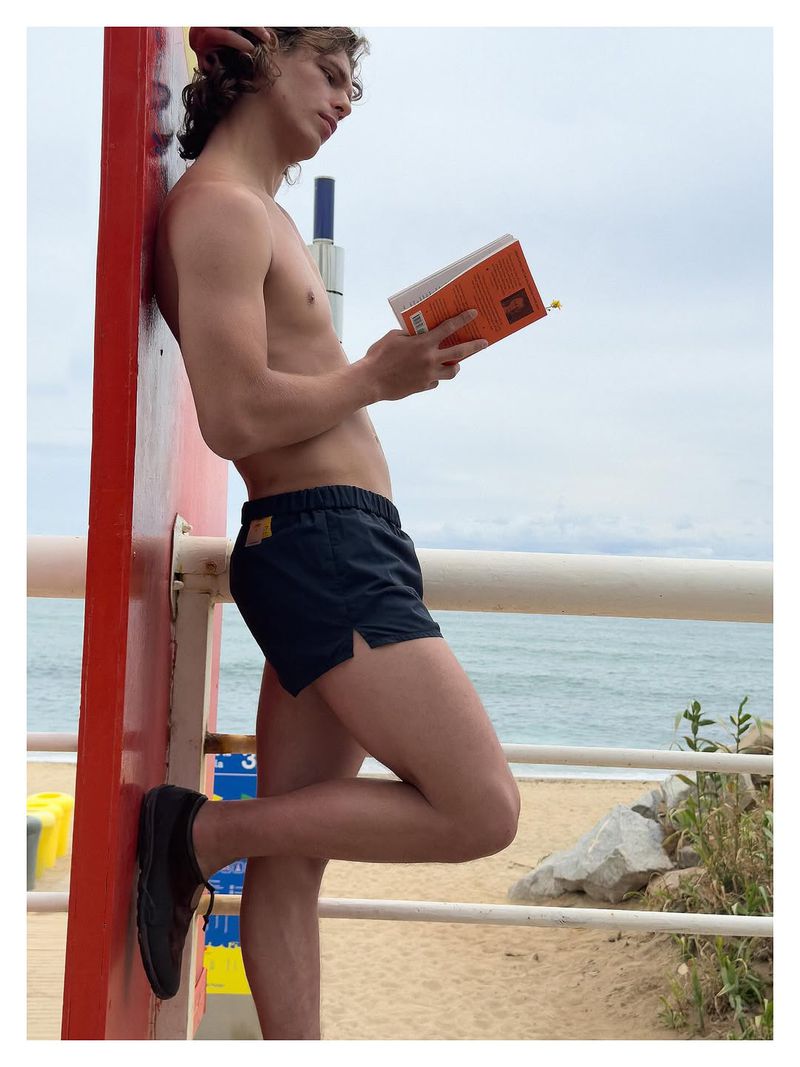 A man in black swim shorts leaning against a red wall and reading an orange paperback book by the beach.