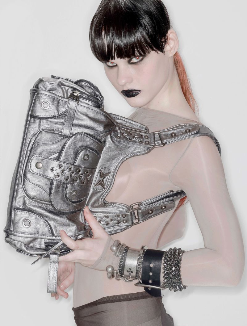 Model with dark eye makeup holding a metallic silver handbag against a white studio background.