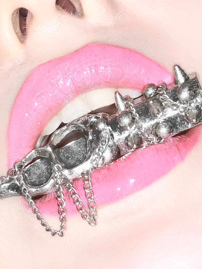 Extreme close-up of glossy pink lips holding an ornate silver spiked ring with hanging chains between white teeth.