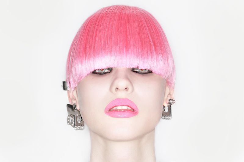 Model Lola Varley with a blunt pink bowl-cut hairstyle, glossy pink lips, and industrial silver earrings on a white seamless backdrop.