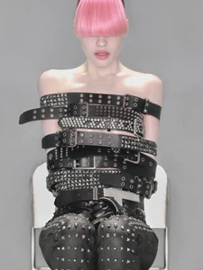 Model with vibrant pink bangs covering her eyes, wearing stacked black studded belts around her torso against a grey backdrop.