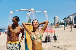 Man and smiling woman standing under an outdoor beach shower next to a surfboard on a sunny sandy beach.
