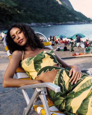 Woman in a green and yellow tropical leaf print two-piece set lounging on a beach chair overlooking a crowded coast.
