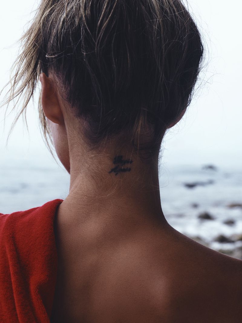 Close-up of Grace Elizabeth from behind showing a small neck tattoo and red towel with an ocean backdrop.