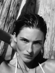 Close-up black and white portrait of model Grace Elizabeth with slicked-back hair posing against weathered wooden posts.