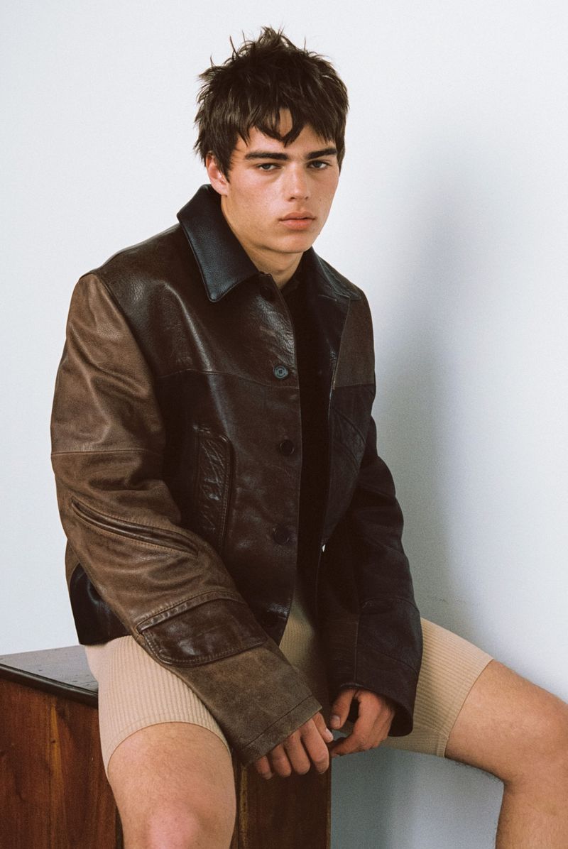 Young man in a distressed two-tone brown leather jacket and beige ribbed shorts seated against a plain white wall.