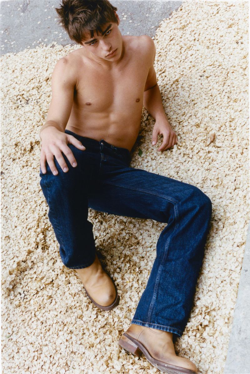 Shirtless young man reclining in dark denim jeans and tan leather boots on a bed of pale wood shavings.