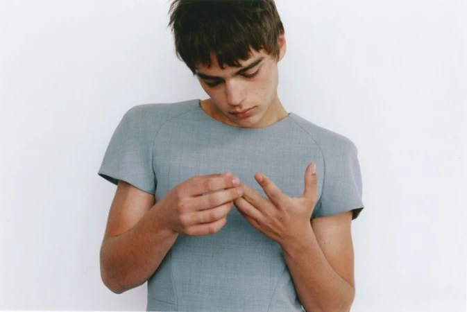 Young man looking down at his hands wearing a structured light grey short-sleeve top against a plain white studio background.