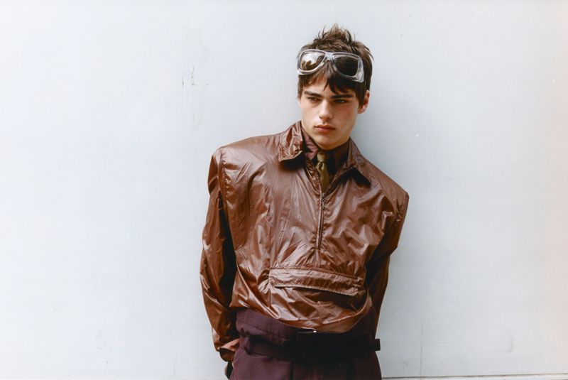 Young man wearing a glossy brown windbreaker and clear goggles on his head against a white wall.