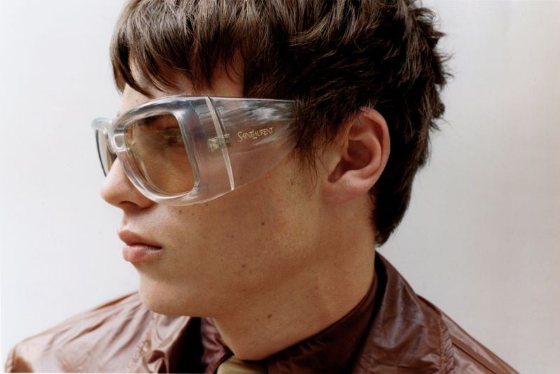 Close-up profile of a man wearing clear shield sunglasses and a glossy brown jacket against a white studio background.