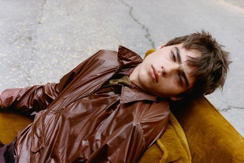 Young man in a shiny brown jacket reclining his head back on a mustard velvet armchair outdoors on cracked asphalt pavement.