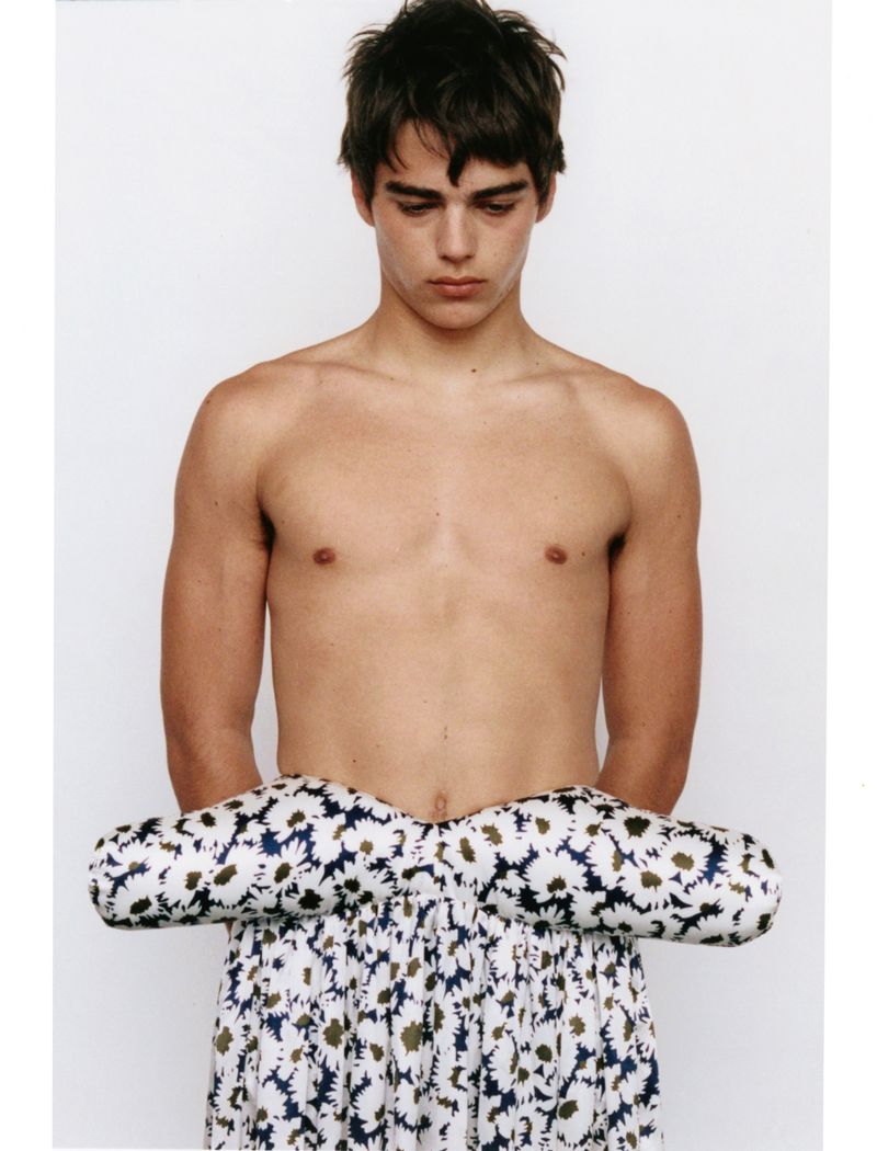 Shirtless young man wearing a padded patterned floral skirt against a clean white studio background.