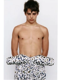 Shirtless young man wearing a padded patterned floral skirt against a clean white studio background.