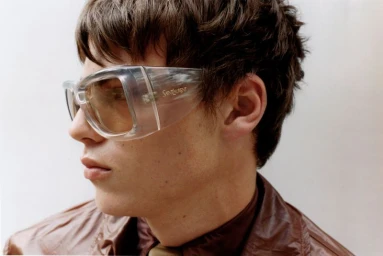 Close-up profile of a man wearing clear shield sunglasses and a glossy brown jacket against a white studio background.