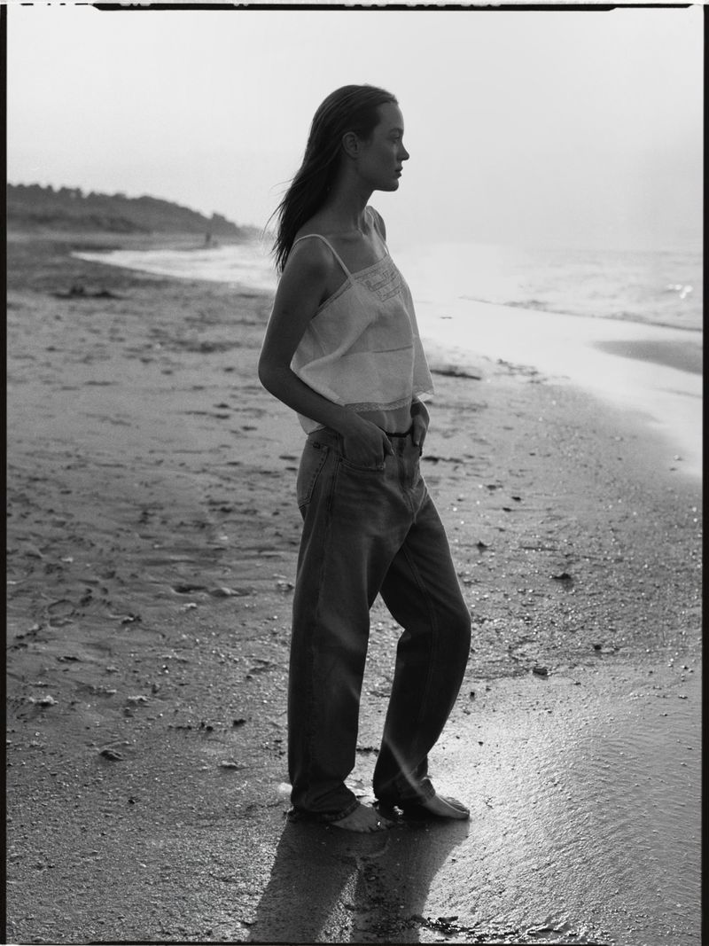 Woman standing on a sandy beach wearing a white tank top and denim jeans in a black and white editorial photograph.