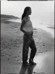 Woman standing on a sandy beach wearing a white tank top and denim jeans in a black and white editorial photograph.