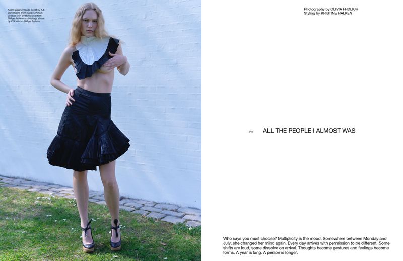 Fashion editorial shot: Blonde woman poses in a vintage-inspired top and skirt on a grassy lawn against a white wall on a sunny day.