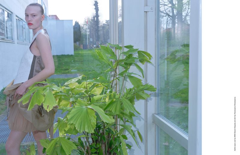 Fashion image featuring a slender adult female posing in a sleeveless top and skirt, next to a potted plant by a window with outdoor views.