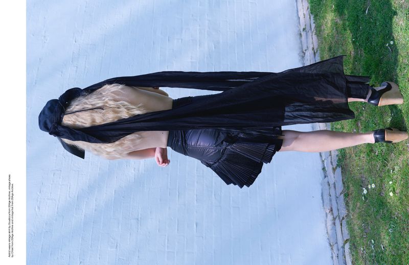 Woman in black dress with blonde hair poses outdoors against a light blue brick wall with green grass, wearing tan platform shoes.