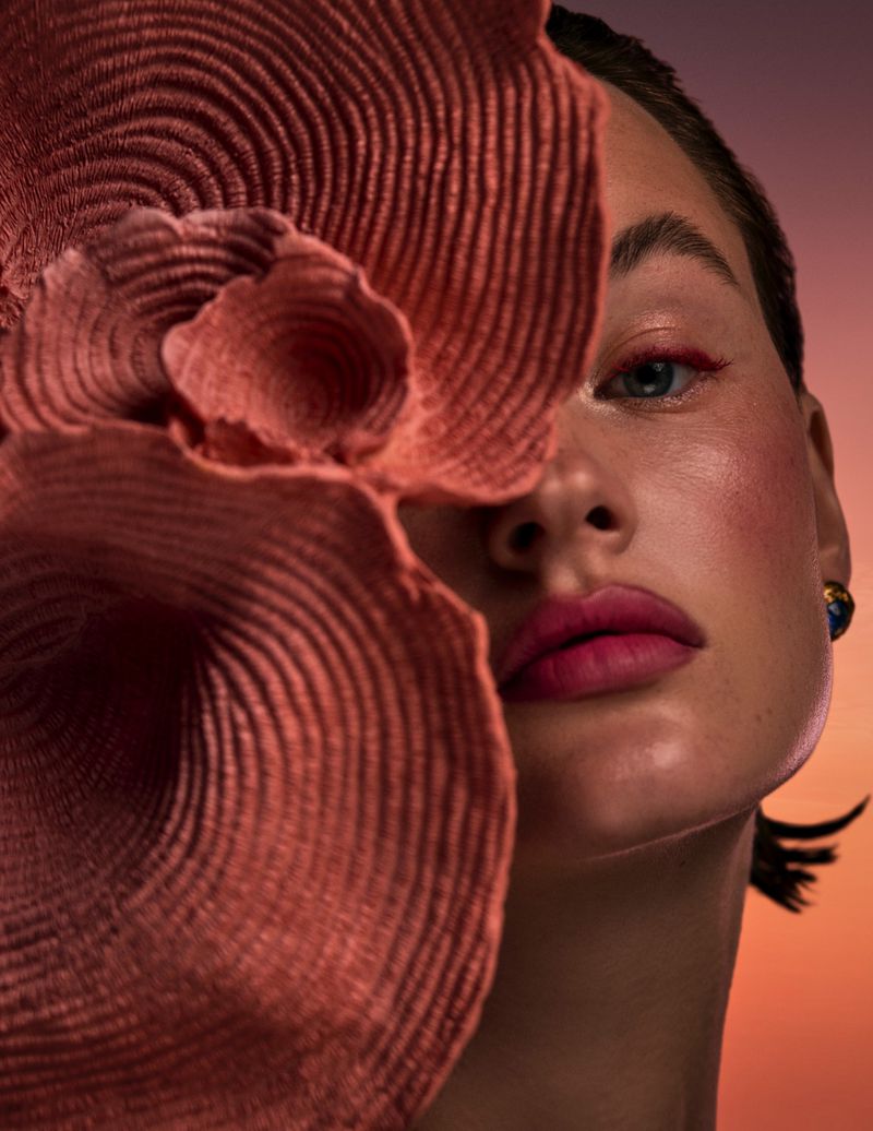 Close-up beauty portrait of a woman partially obscured by a pleated coral woven brim against a warm sunset gradient background.