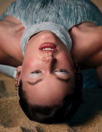 Close-up portrait of a woman lying upside down on sand wearing a textured pale blue halter top and blue eyeshadow.