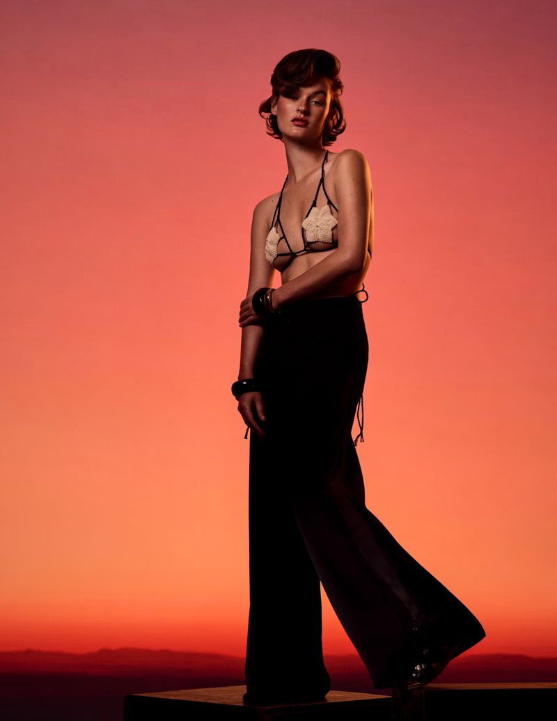 Model wearing a sheer applique bralette and high-waisted black wide-leg trousers posing against a vibrant orange and red sunset sky.