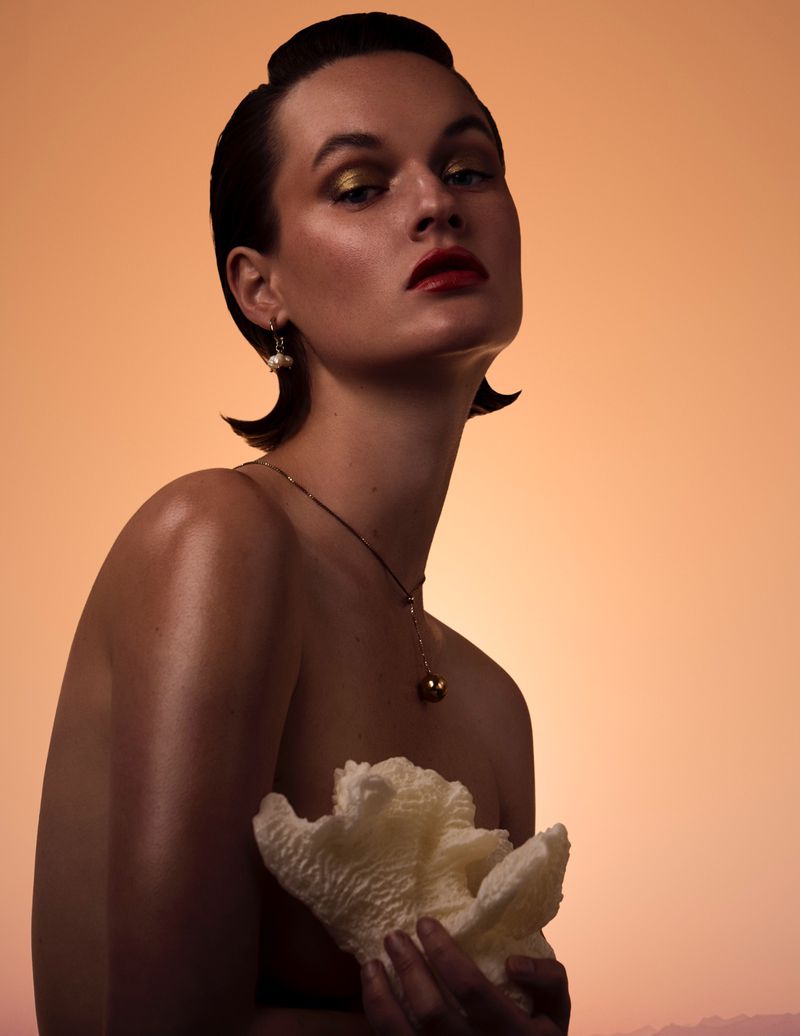 Beauty portrait of a woman with slicked-back hair holding a white coral piece against a warm peach gradient background.