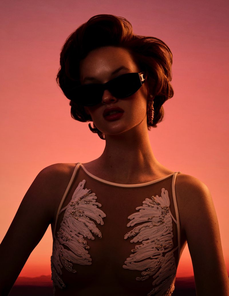 Woman in black sunglasses and a white embroidered sheer dress posed against a glowing orange and pink sunset sky.