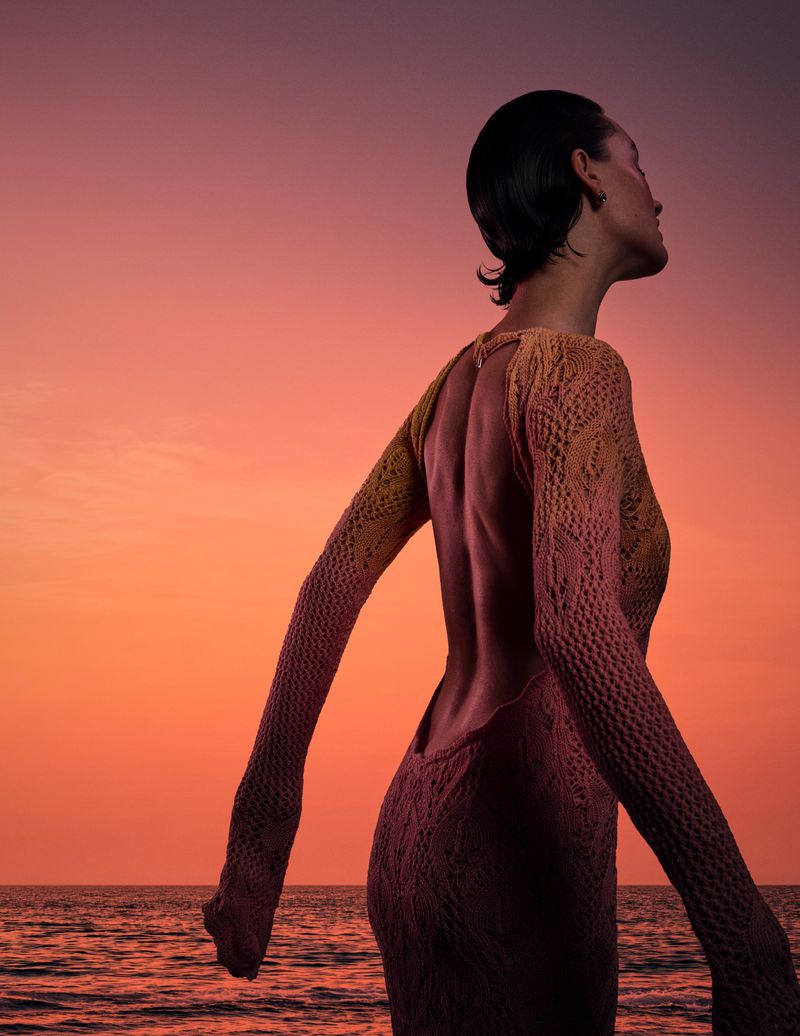 Model wearing an open-back knitted dress looking away against an orange sunset ocean sky.