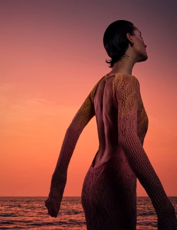 Model wearing an open-back knitted dress looking away against an orange sunset ocean sky.