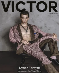 Male model Ryder Forsyth seated on a chair wearing a patterned pink and brown suit jacket and matching trousers against a textured grey background.