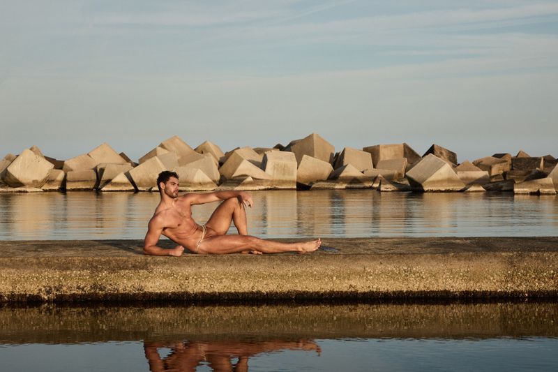 A man reposes on a concrete pier by calm water against a backdrop of stacked rock formations under an expansive sky.