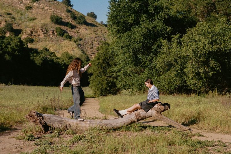 Two models in casual designer outfits pose on a large fallen tree trunk in a sunlit grassy field bordered by green trees.