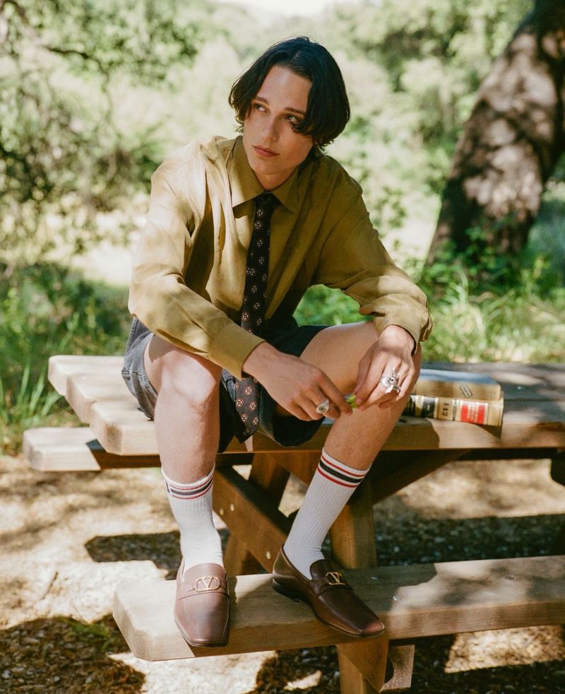 Model Etienne Maclaine sitting on an outdoor wooden picnic bench wearing a khaki button-down shirt, necktie, shorts, and brown loafers.
