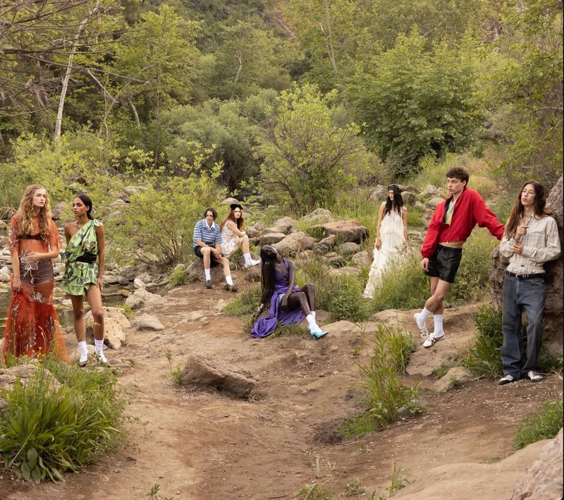 Group of fashion models posing outdoors along a rocky dirt path and stream surrounded by lush green trees for L'Officiel USA.
