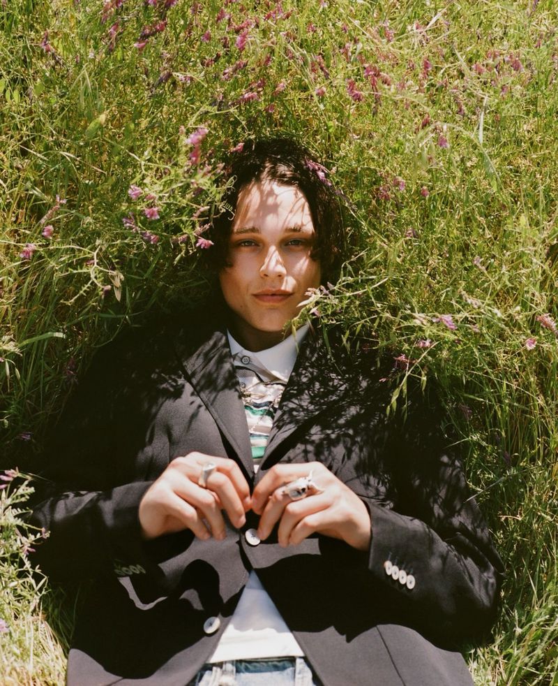 Model Etienne Maclaine lying in tall grass and wildflowers wearing a black tailored blazer for L'Officiel USA.