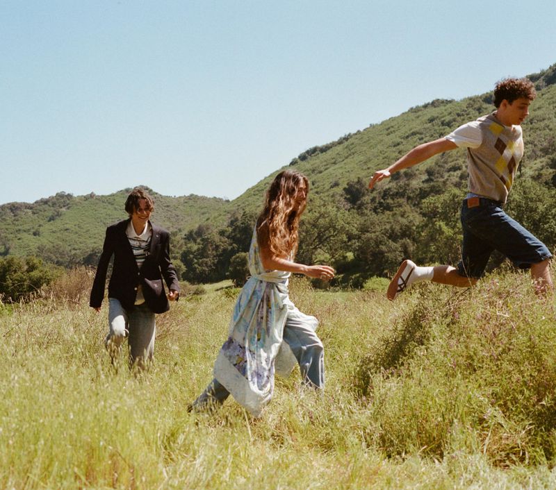 Three models running across a sunlit grassy field in front of rolling green hills in an outdoor editorial scene.