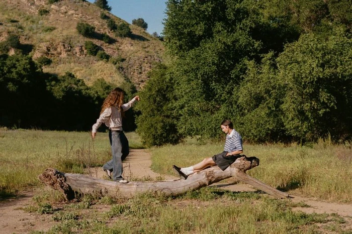 Two models in casual designer outfits pose on a large fallen tree trunk in a sunlit grassy field bordered by green trees.