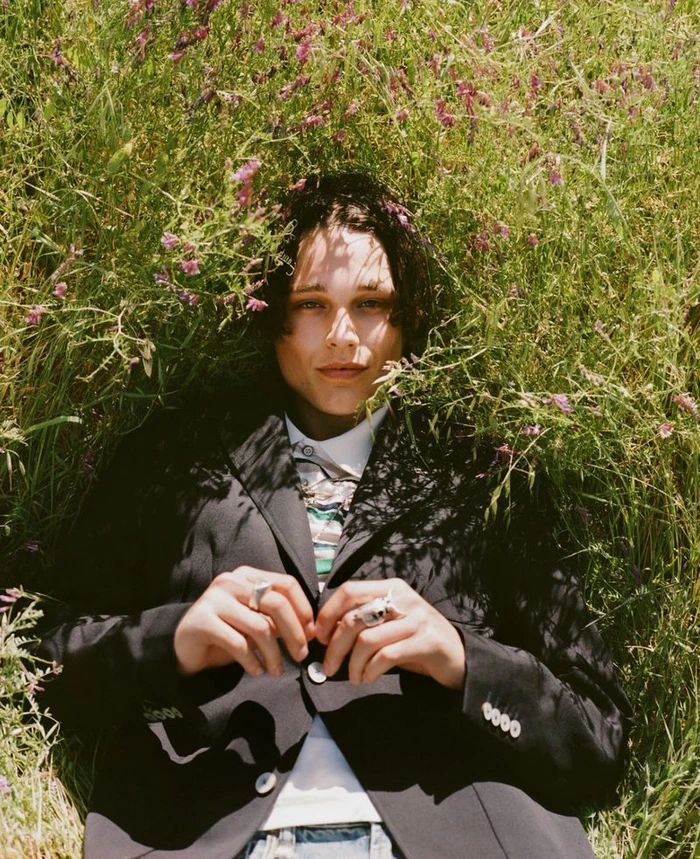 Model Etienne Maclaine lying in tall grass and wildflowers wearing a black tailored blazer for L'Officiel USA.
