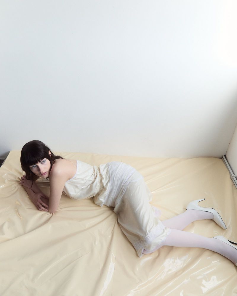Model lying on a glossy cream plastic surface wearing a light-coloured dress and white high heels.