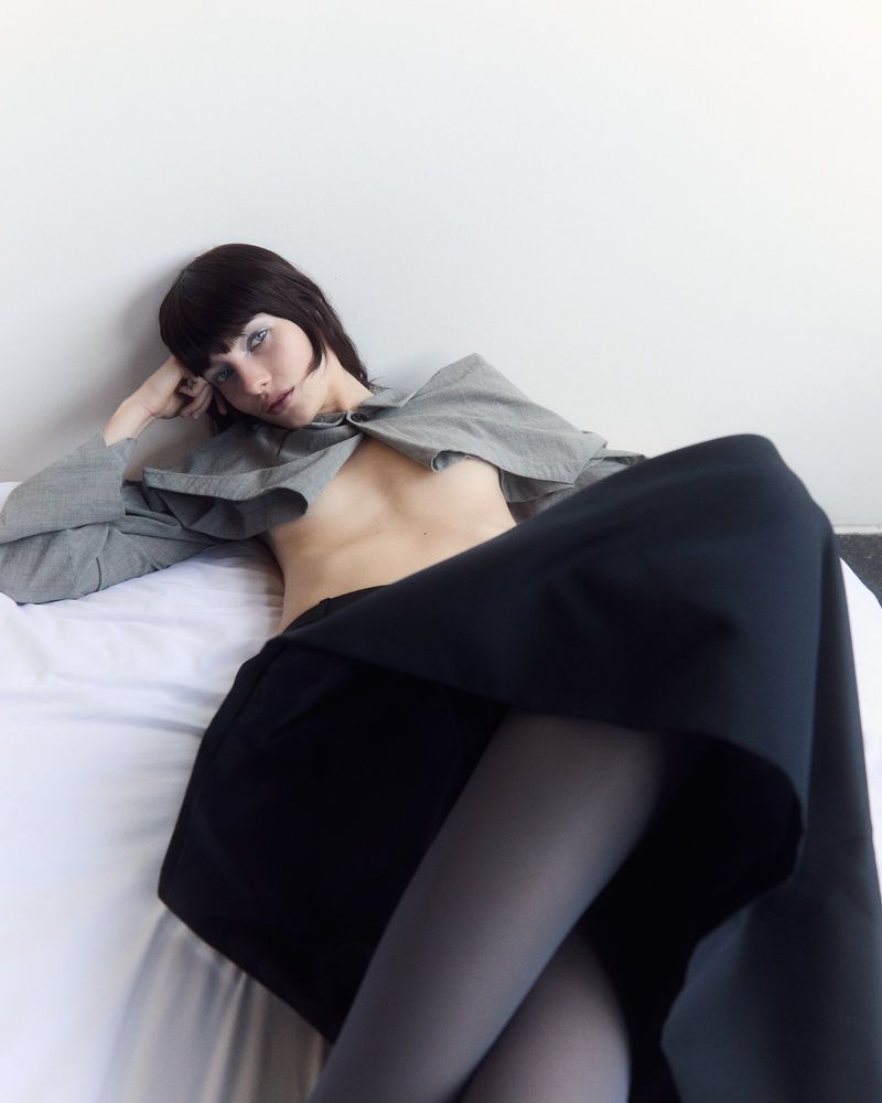 Model Clara Denison reclining in an open grey cropped jacket and black skirt on white bedding against a white wall.